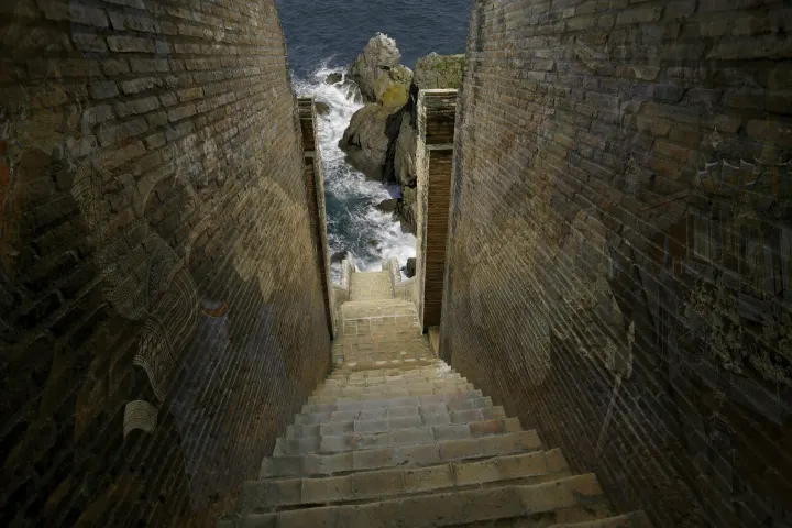 Escalier de briques descendant entre deux hauts murs vers des rochers battus par la mer, avec des motifs architecturaux en transparence.